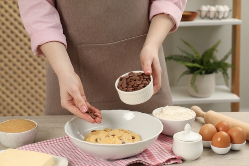 a woman in an apron sprinkles chocolate chips from a measuring cup into a bowl