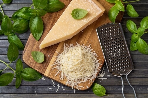 a wedge of parmesan cheese sits on a cutting board next to a pile of cheese shavings. basil leaves are sprinkled over top